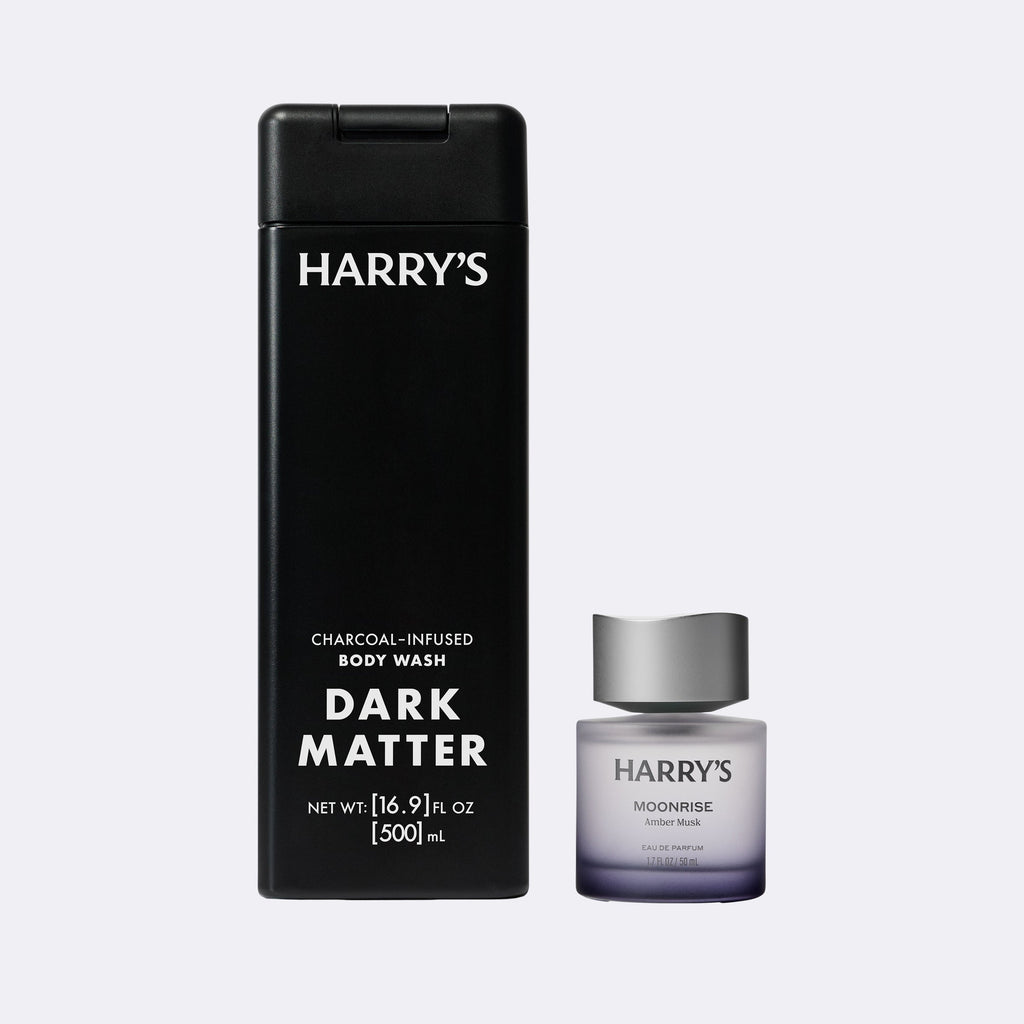 Harry's Dark Matter charcoal-infused body wash shown with its tall black bottle and a smaller Moonrise cologne bottle on a light gray background.