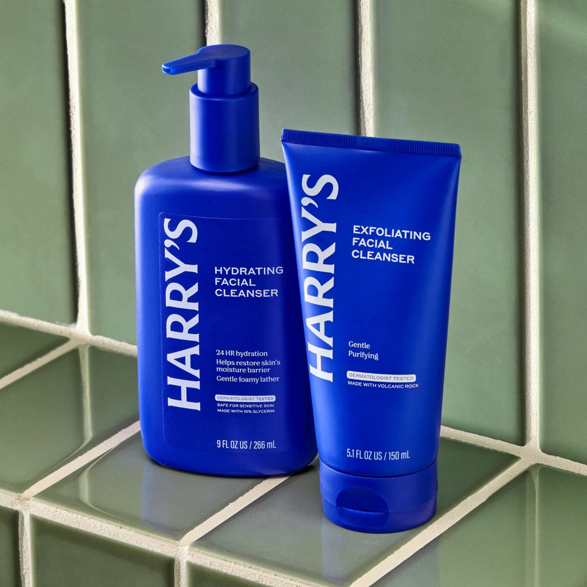 Two blue bottles of Harry's facial cleanser on a green tiled surface