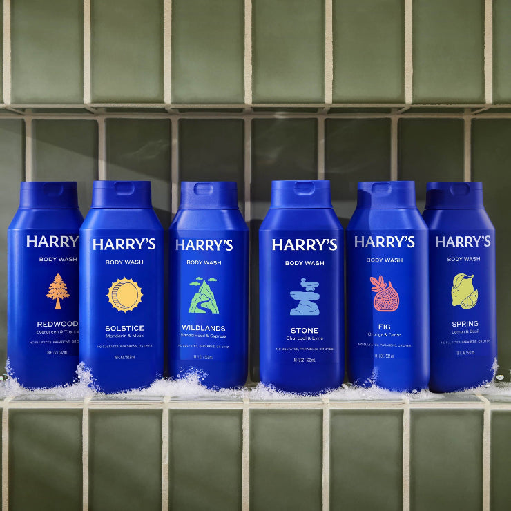 Six bottles of Harry's body wash with different scents on a tiled surface.