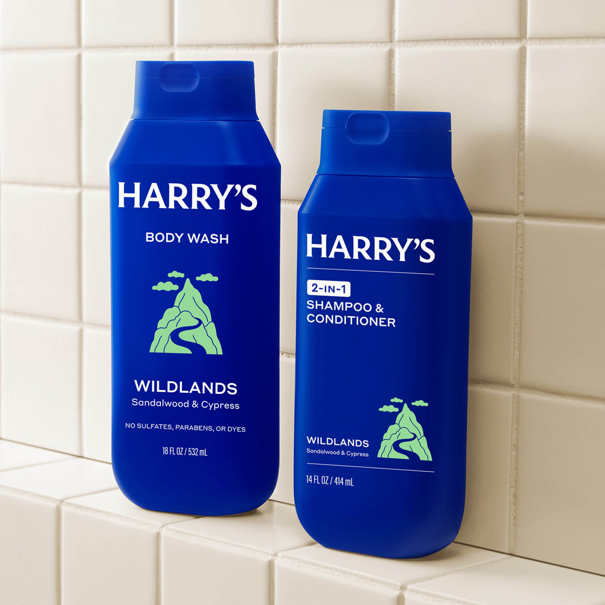 Two blue bottles of Harry's Wildlands body wash and shampoo & conditioner on a tiled shelf.
