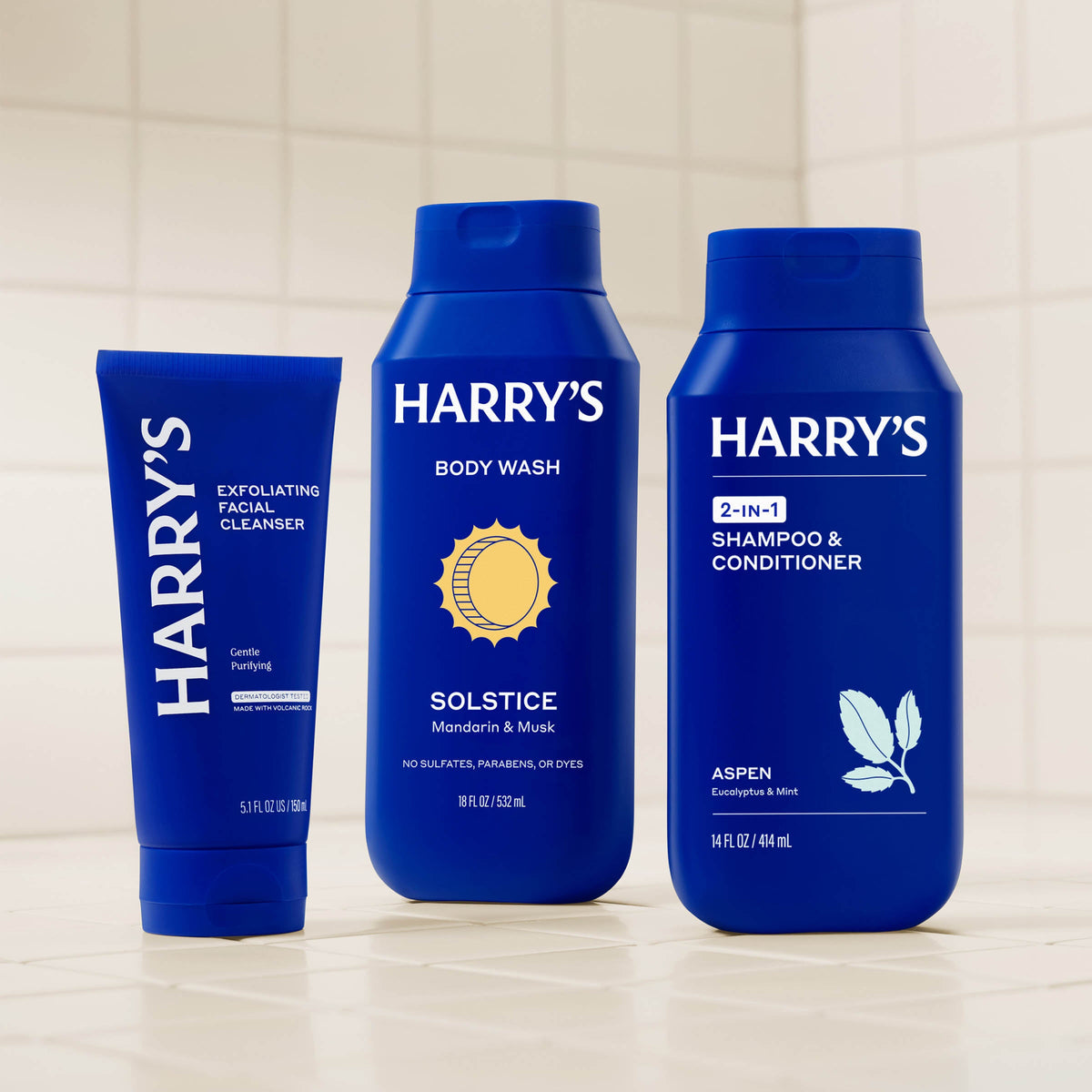 Three blue bottles of Harry's grooming products on a tiled bathroom wall background.