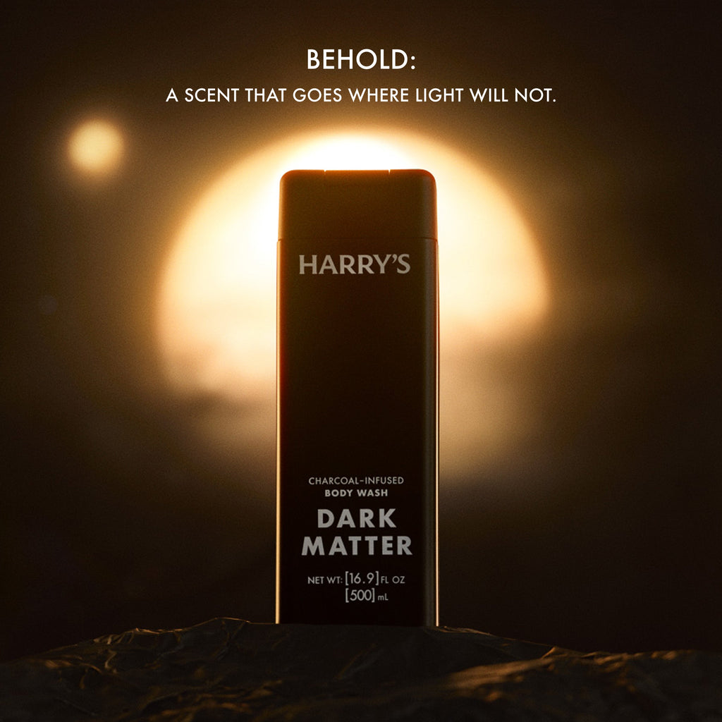 A single rectangular body wash bottle labeled HARRY'S Dark Matter Charcoal Infused Body Wash stands upright on a dark textured surface against a dramatic glowing orange and black background. White text at the top reads, 'BEHOLD: A SCENT THAT GOES WHERE LIGHT WILL NOT.