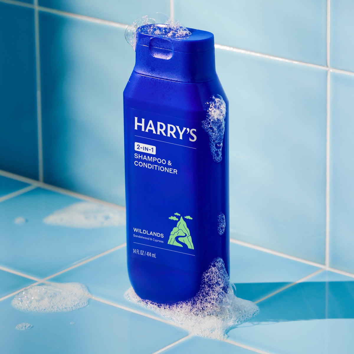 Blue bottle of 'Harry's' shampoo and conditioner on a tiled bathroom floor with water droplets.