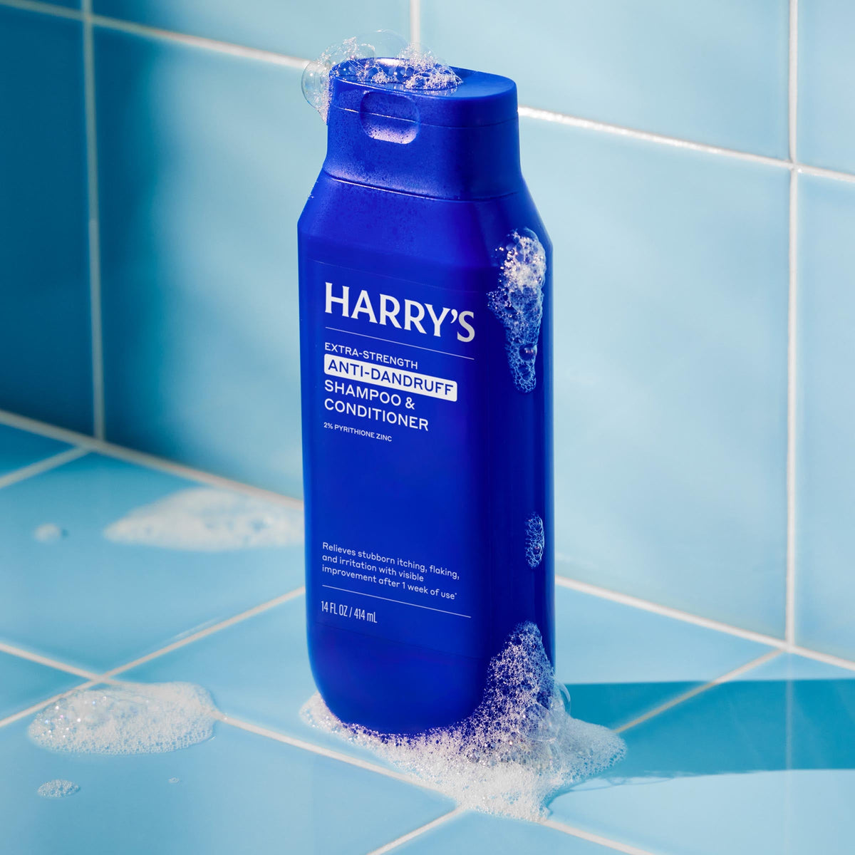 Blue bottle of Harry's shampoo and conditioner on a tiled surface with water droplets.