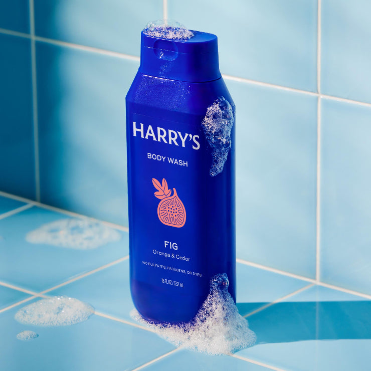 Blue bottle of Harry's body wash with fig scent on a tiled bathroom shelf.