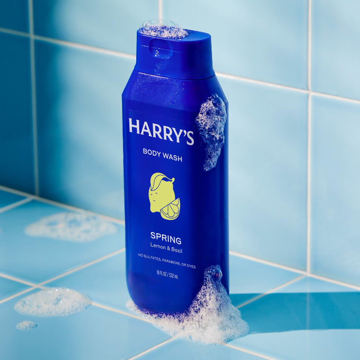Blue bottle of Harry's body wash on a tiled bathroom shelf with soap suds.