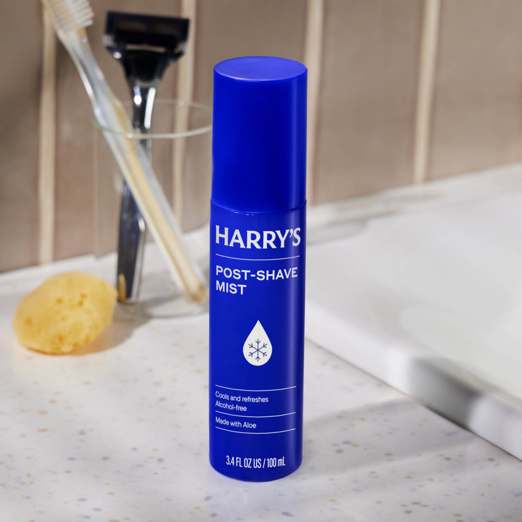 Blue bottle of Harry's post-shave mist on a bathroom counter with a razor and sponge in the background.