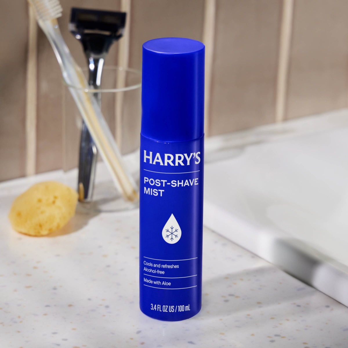 Blue bottle of Harry's post-shave mist on a bathroom counter with a razor and sponge in the background.