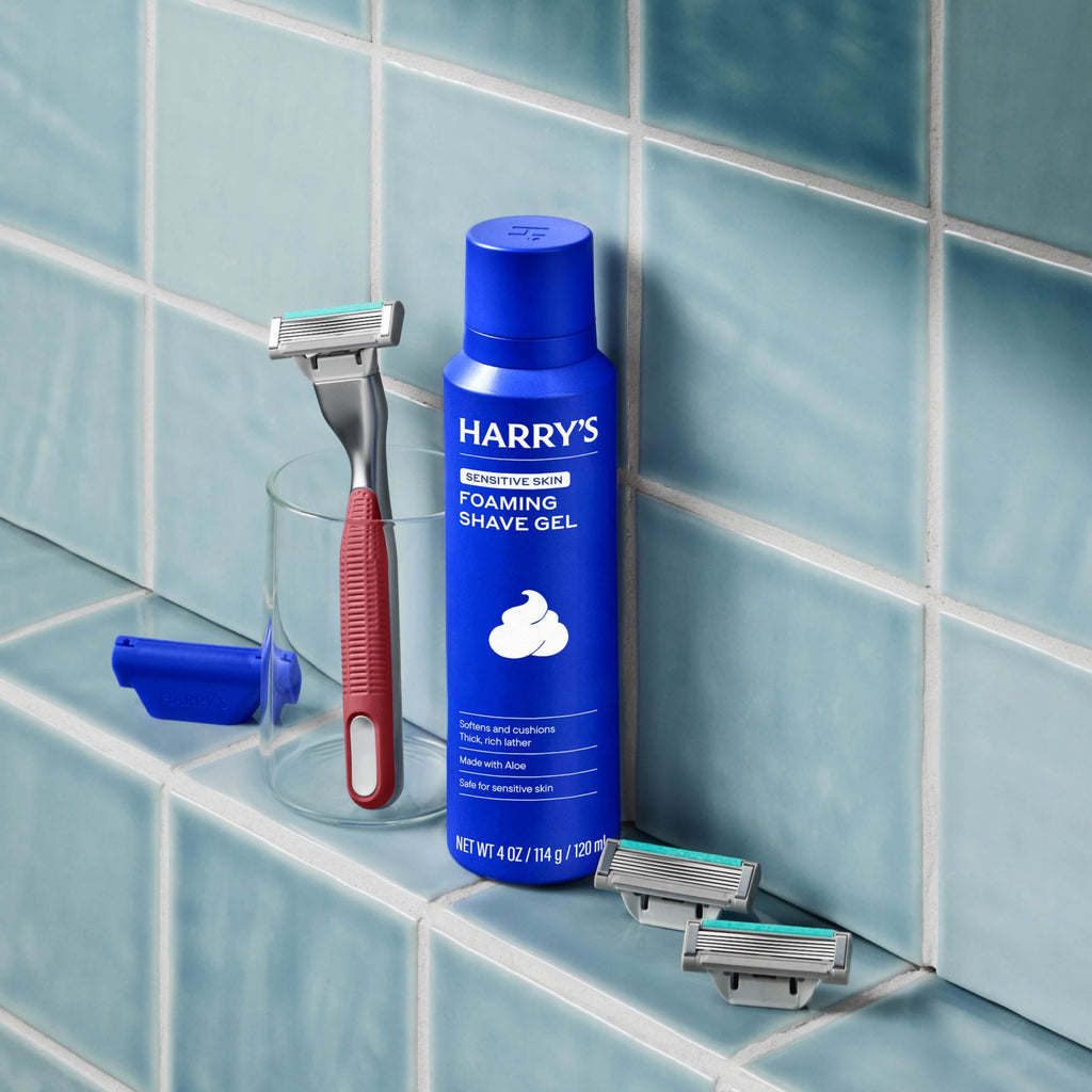 Harry's foaming shave gel bottle with a razor and razors on a tiled bathroom shelf.