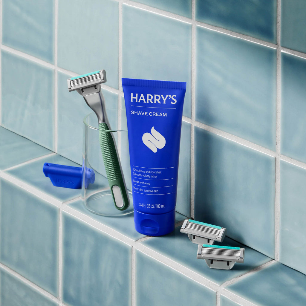 Harry's shave cream tube with razors on a tiled bathroom shelf