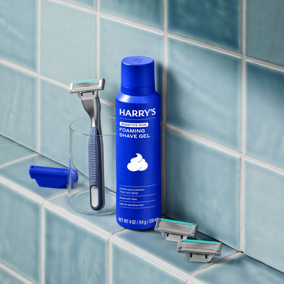 Harry's foaming shave gel bottle with a razor and razor heads on a tiled bathroom shelf.