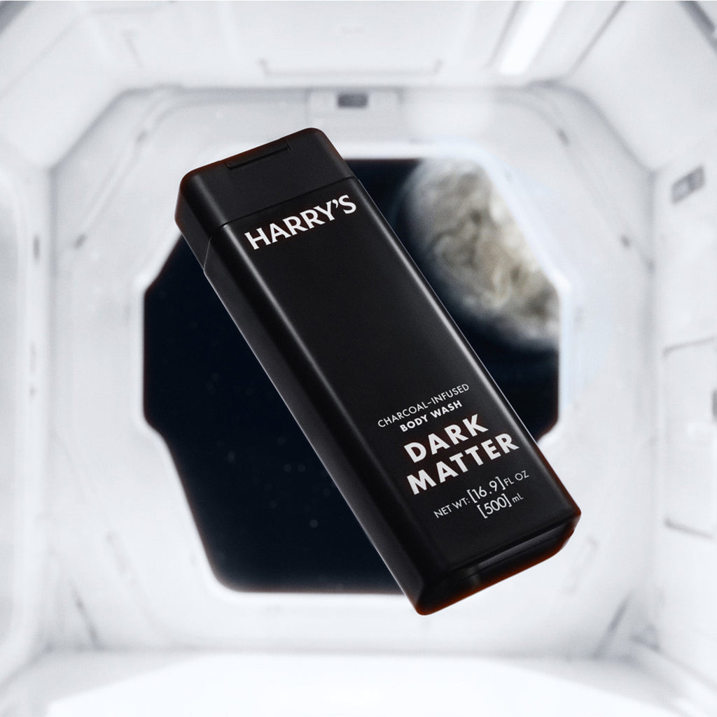A black rectangular bottle of Harry's Dark Matter Charcoal-Infused Foot Wash floating inside a white futuristic spacecraft interior.