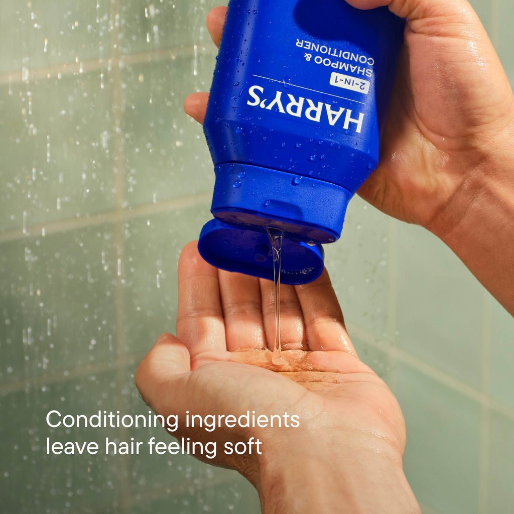 Hand holding a blue bottle of Harrys shampoo with shower in the background