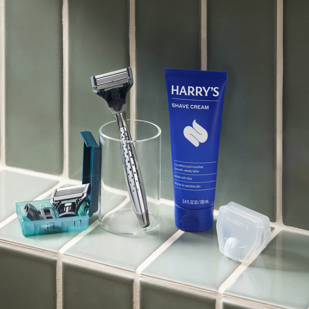 Shaving setup with a razor, glass, and Harry's shave cream on a tiled surface.