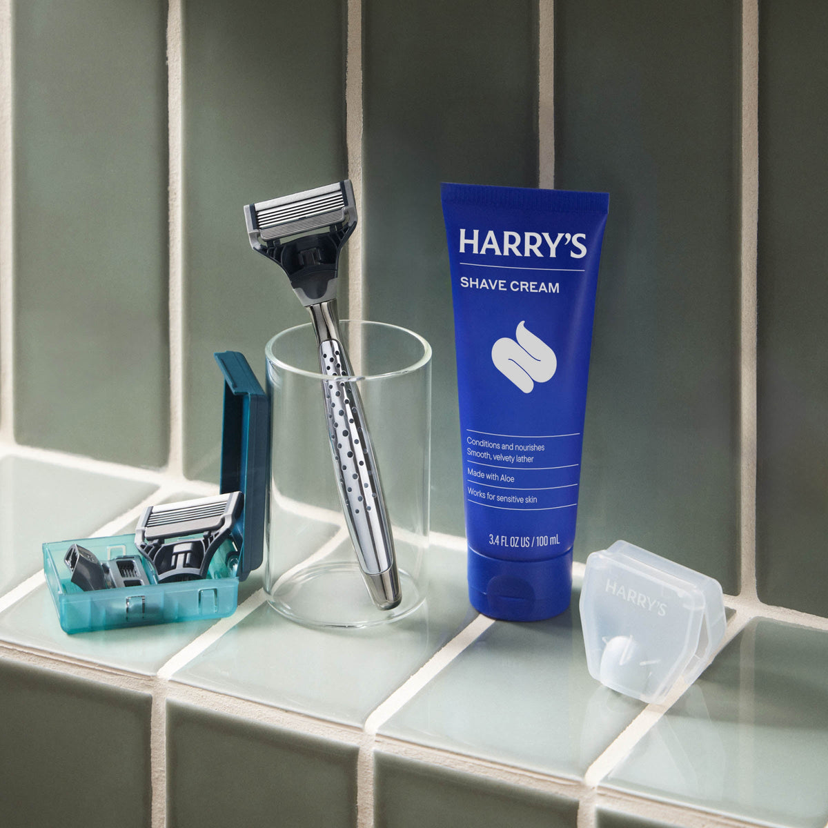 Shaving setup with a razor, glass, and Harry's shave cream on a tiled surface.