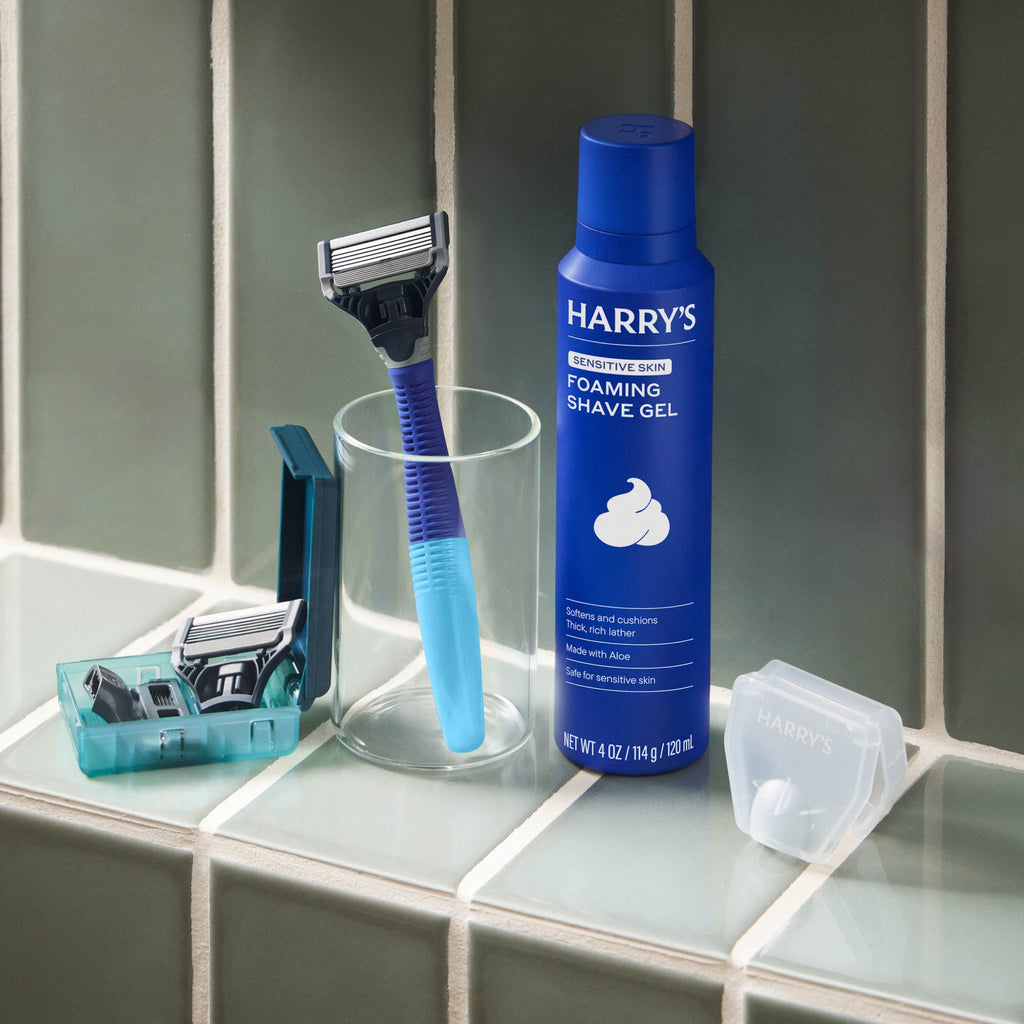 Harry's shaving products including a blue bottle of foaming shave gel, a razor, and razors in a holder on a bathroom counter.