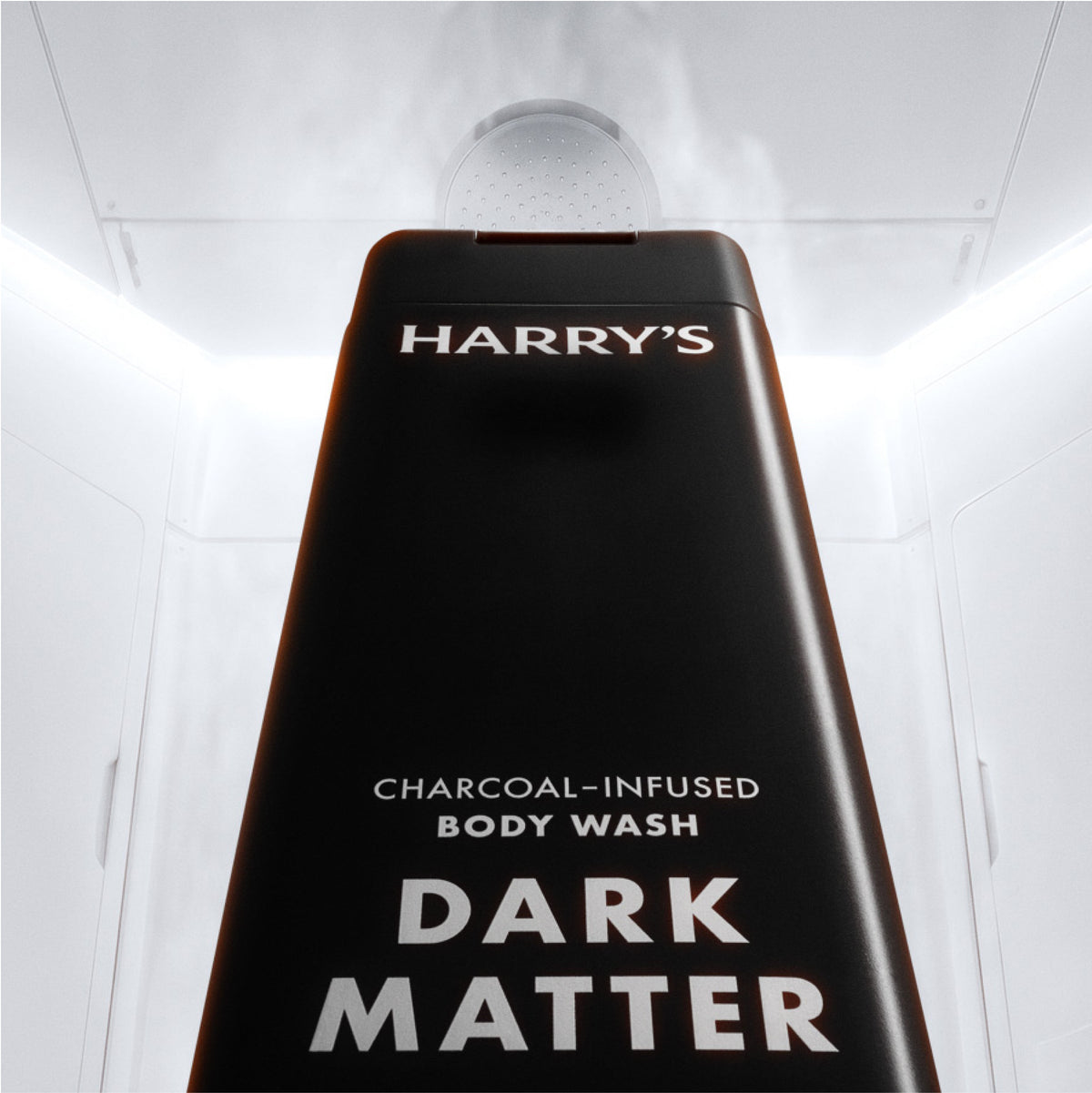 A tall black shower head body wash dispenser labeled HARRY'S CHARCOAL-INFUSED BODY WASH DARK MATTER, shown from a low angle inside a white shower cabin with steam rising from the top.