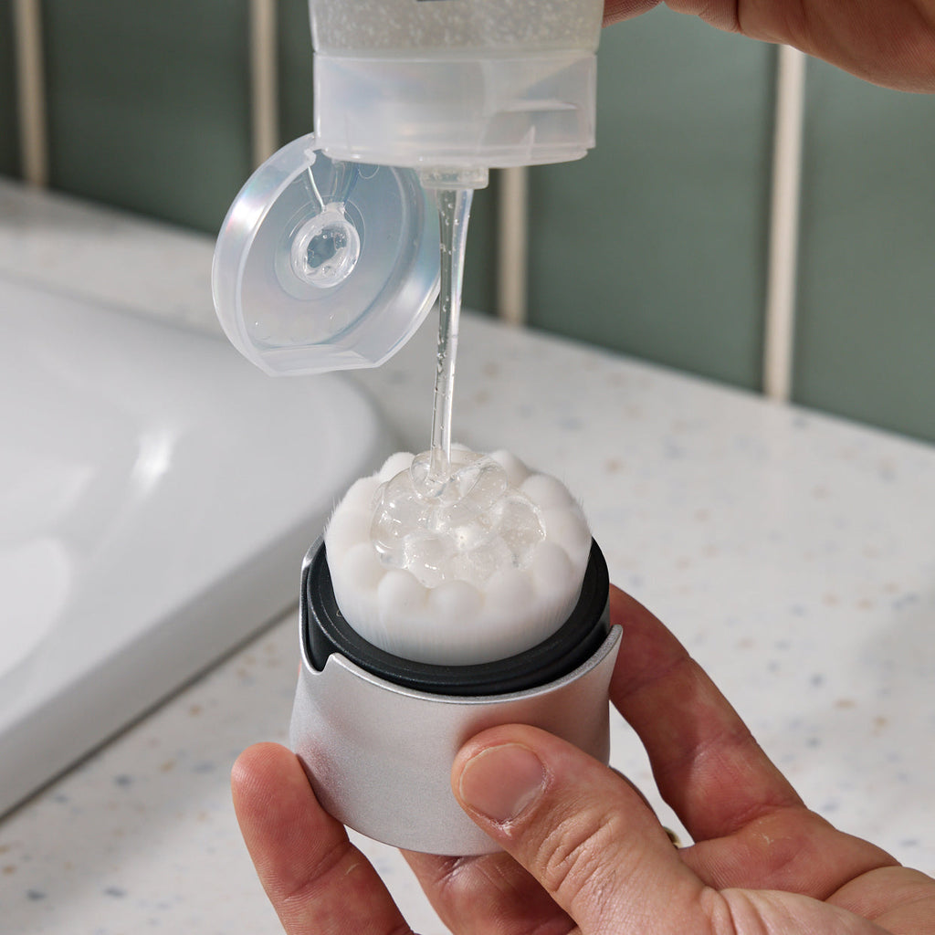 Person using a clear container to pour liquid into a white and black device on a bathroom counter.