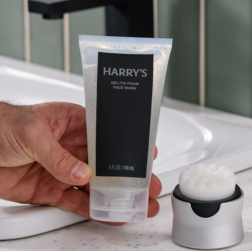 Hand holding a tube of Harry's gel-to-foam face wash with a sink in the background