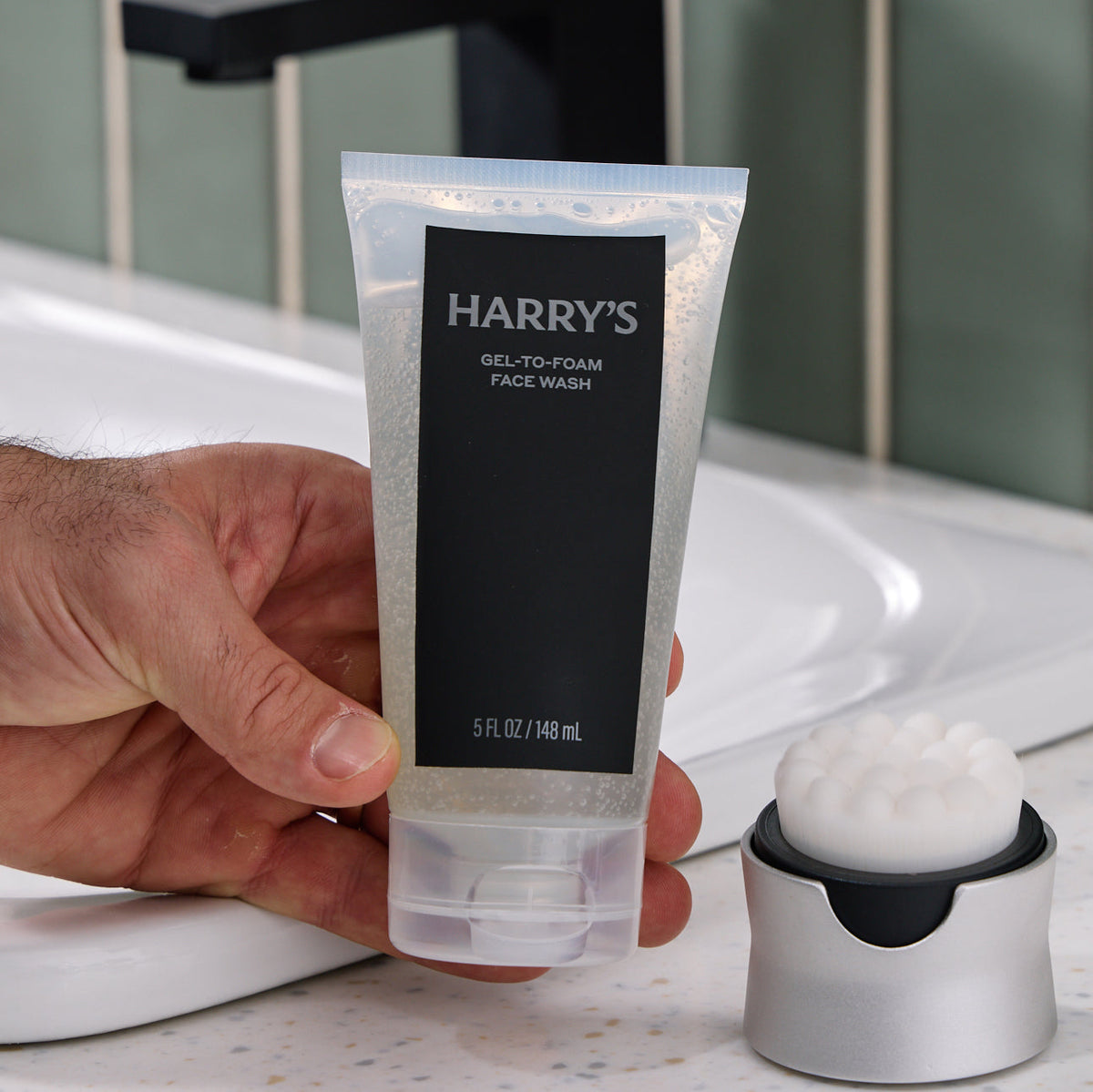 Hand holding a tube of Harry's gel-to-foam face wash with a sink in the background