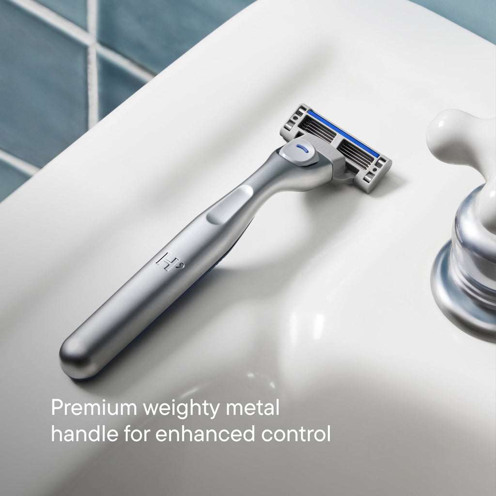 A silver shaving razor with a blue and black multi-cut shaving head, resting on a white ceramic sink next to a chrome faucet. White text at the bottom reads 'Premium weighted metal handle for enhanced control'.