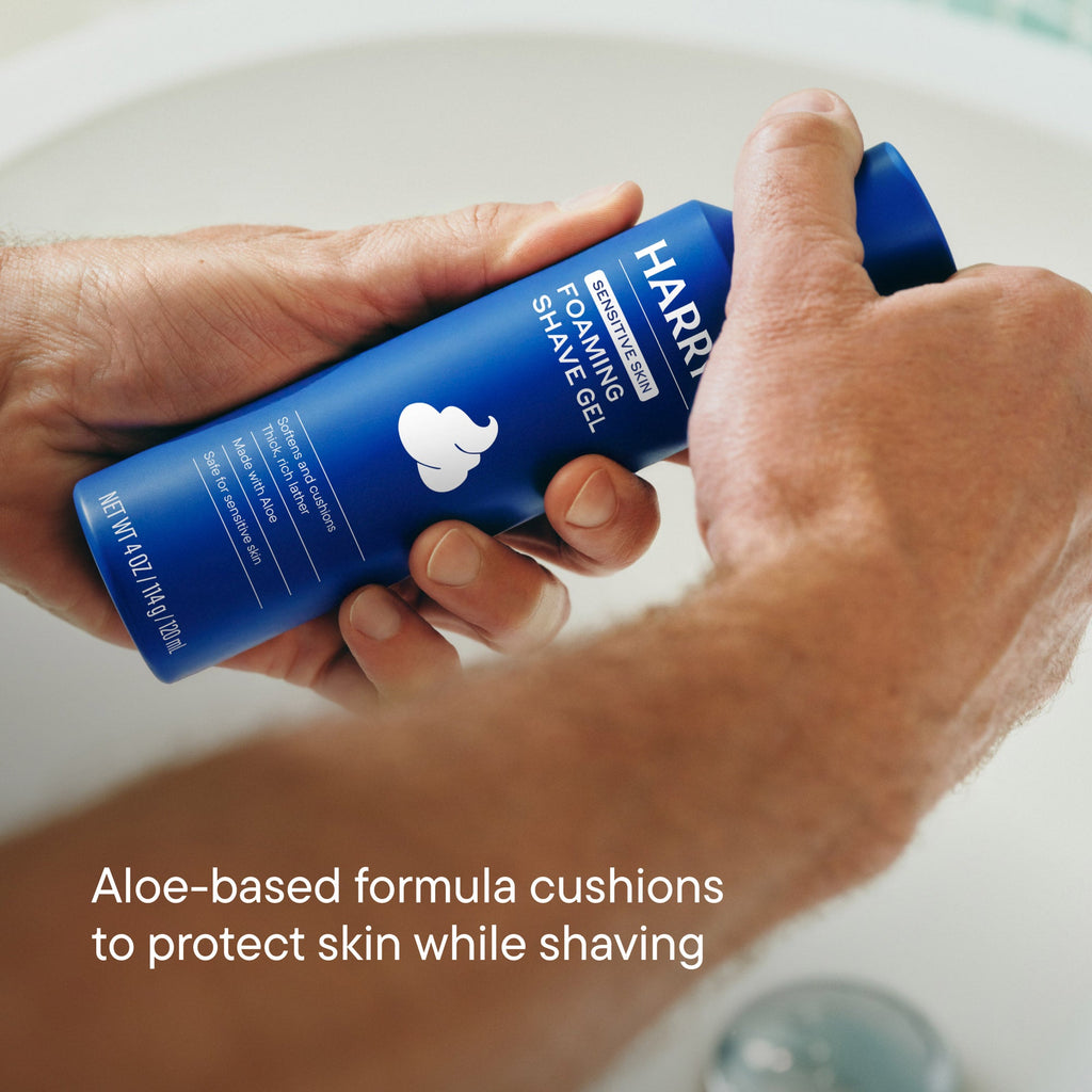 Hand holding a blue bottle of Harry's foaming shave gel with a sink in the background.