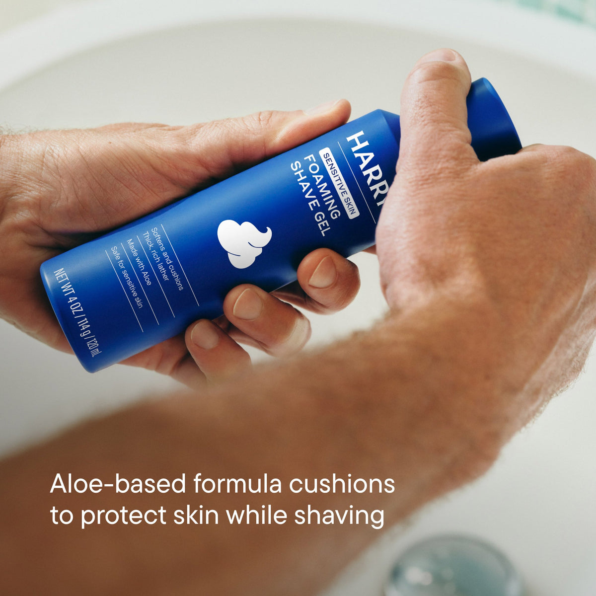 Hand holding a blue bottle of Harry's foaming shave gel with a sink in the background.