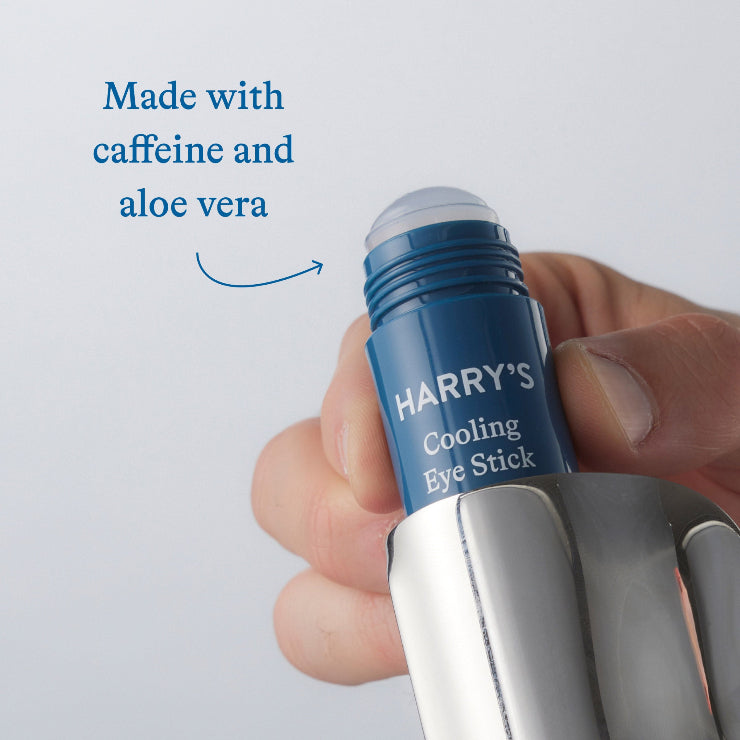 Hand holding a blue 'Harry's' cooling eye stick with text about caffeine and aloe vera on a light gray background.