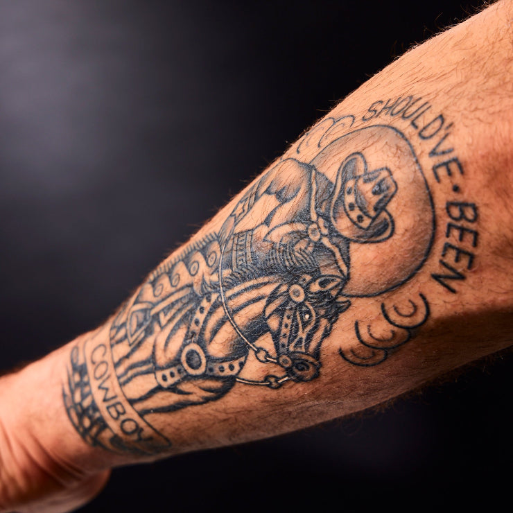 Tattoo of a cowboy on a horse with text on a person's arm against a dark background