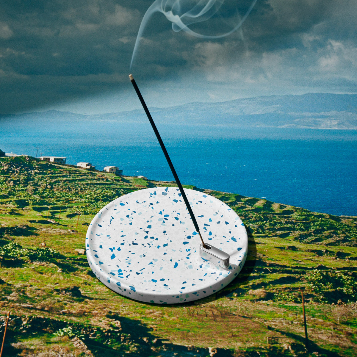 Terrazzo Incense holder with gently burning incense, set over a photo of the Mediterranean sea and countryside.