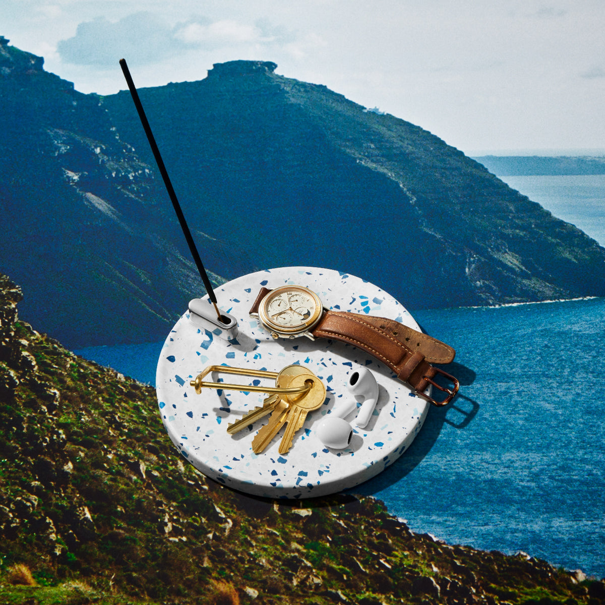 Terrazzo Incense holder with gently burning incense, keys, watch and headphones, set over a photo of the Mediterranean sea and countryside.
