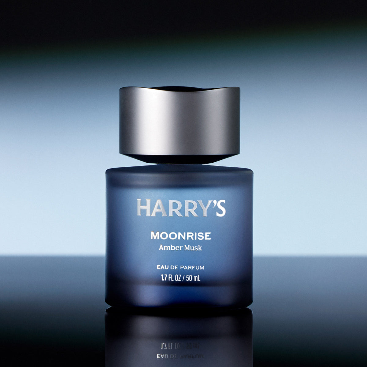 Blue perfume bottle labeled 'Harry's Moonrise Amber Musk' on a reflective surface with a gradient background.