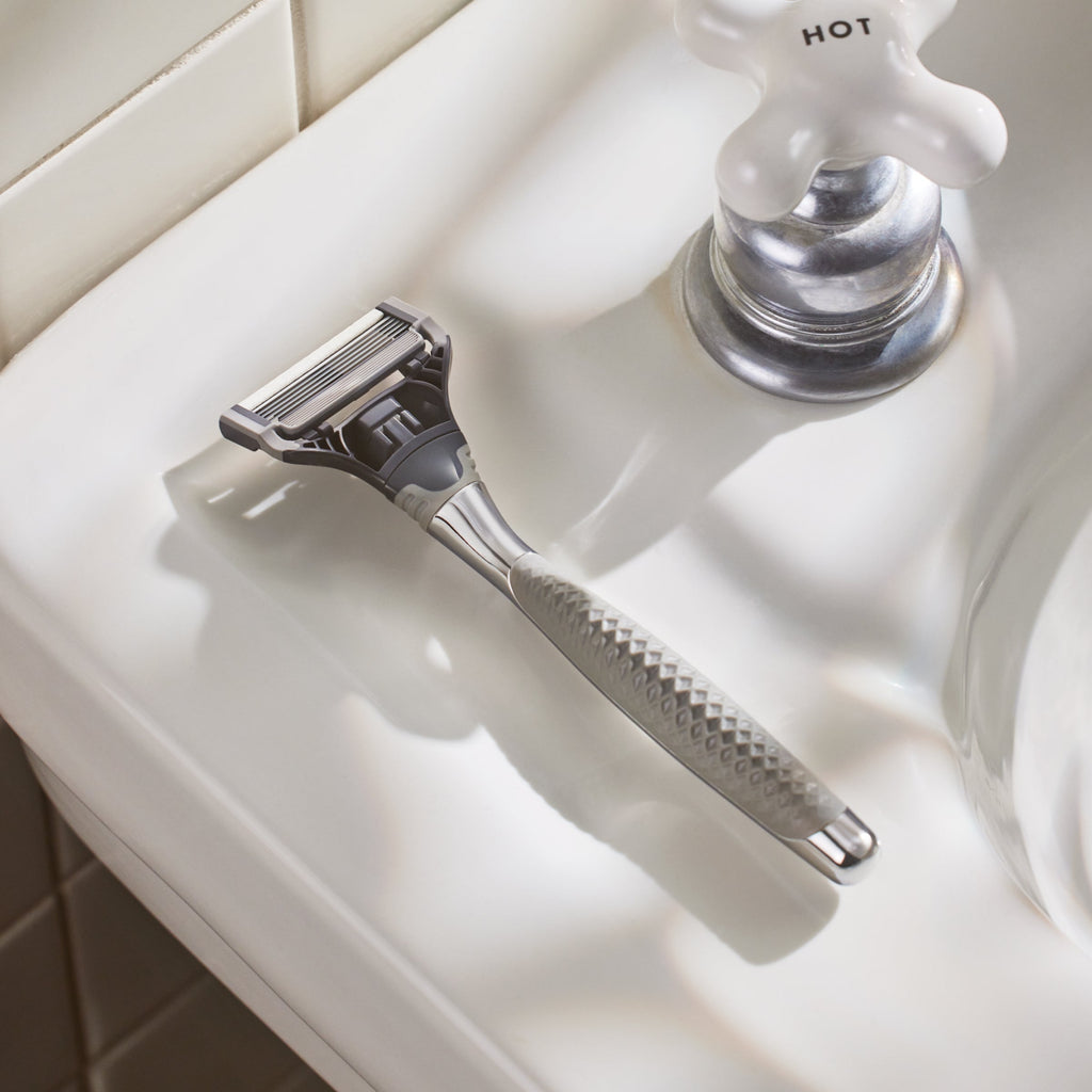 Silver safety razor on a white bathroom counter with a faucet in the background