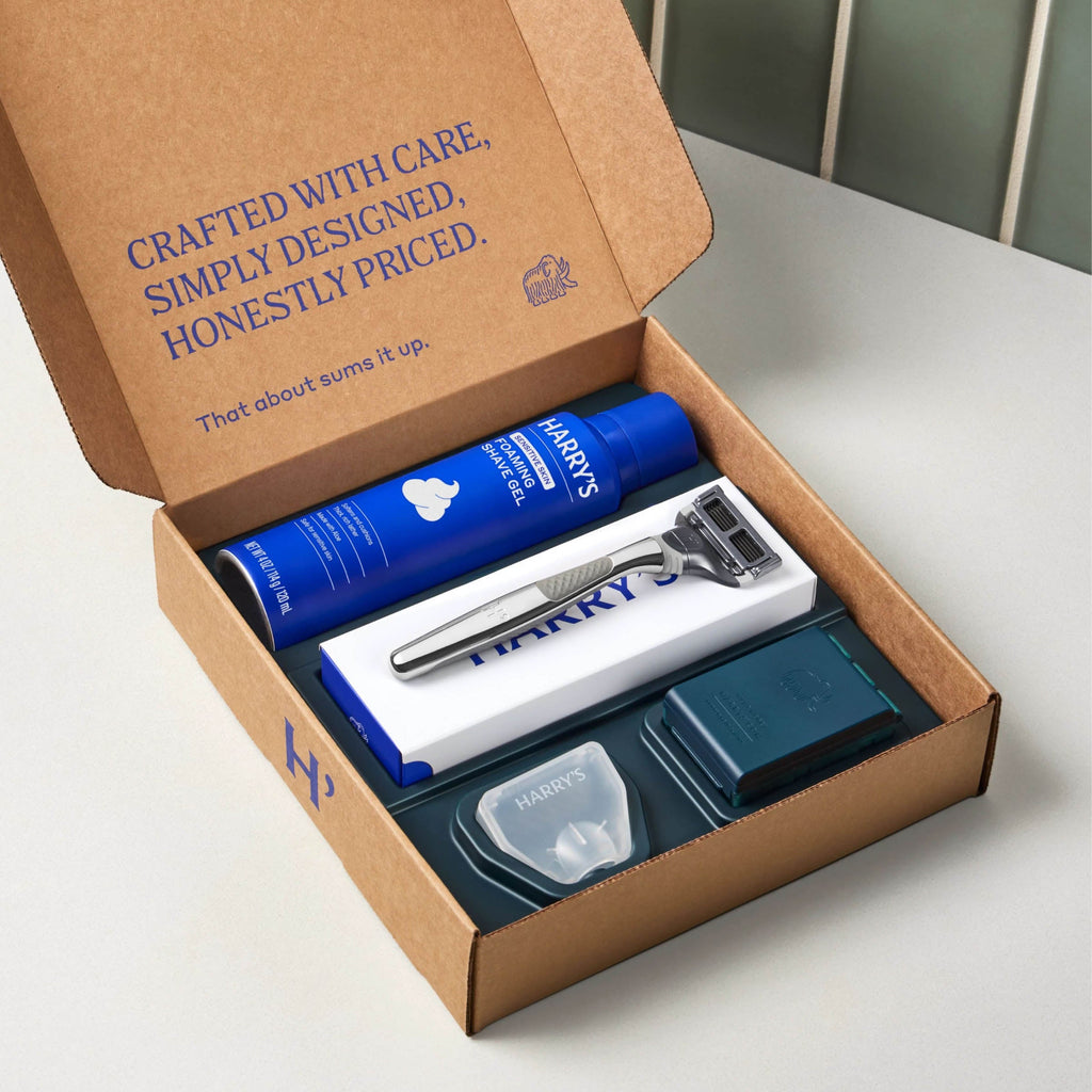 Harry's grooming kit with shaving cream, razor, and other items in a cardboard box on a white surface.