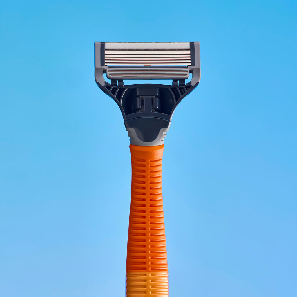 A close-up studio shot of a razor head and part of its handle against a solid light blue background. The product features a dark gray plastic head with metallic silver blades and an orange textured handle.