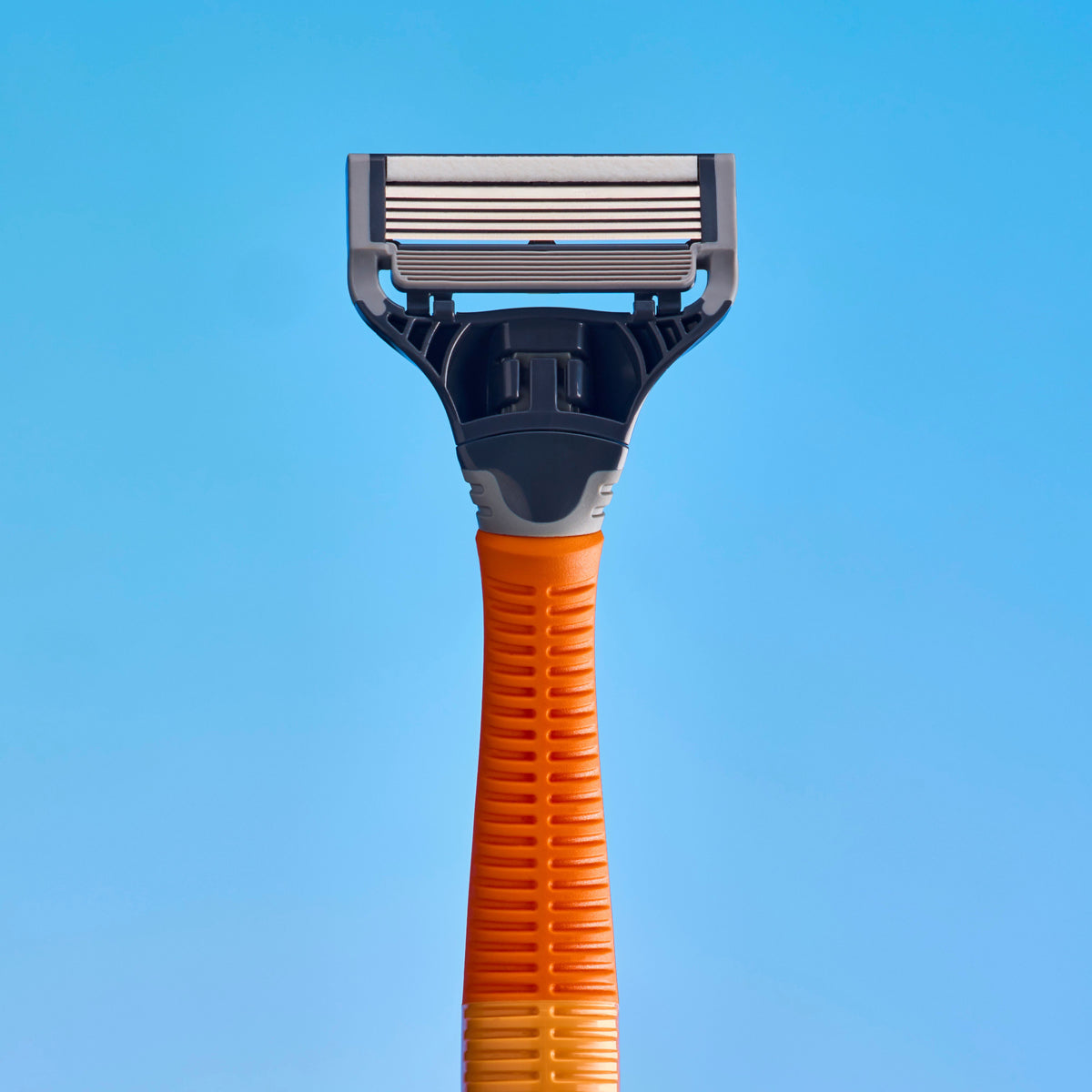 A close-up studio shot of a razor head and part of its handle against a solid light blue background. The product features a dark gray plastic head with metallic silver blades and an orange textured handle.