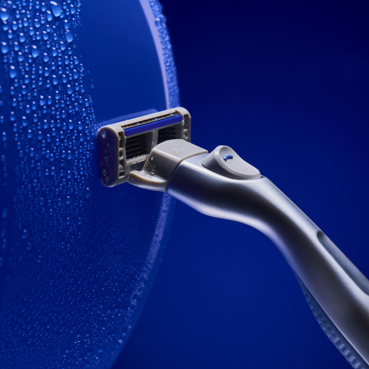A close-up studio shot of a metallic silver electric razor with a blue accent on the head, positioned against a dark blue background with water droplets on a curved surface.