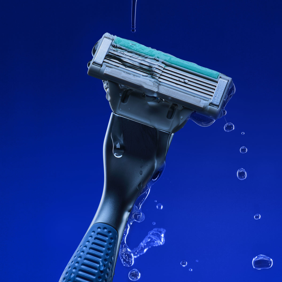 A close-up studio shot of a razor with a teal and gray blade, shown with water droplets and splashes against a vivid blue background.